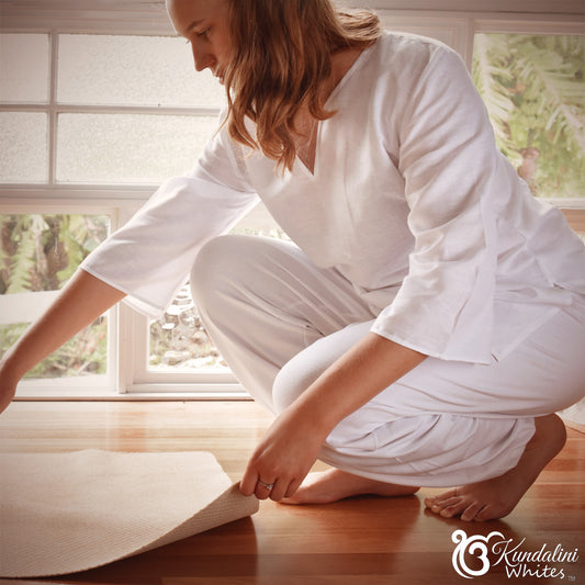 Woman in white outfit preparing a yoga mat in a bright room with windows.