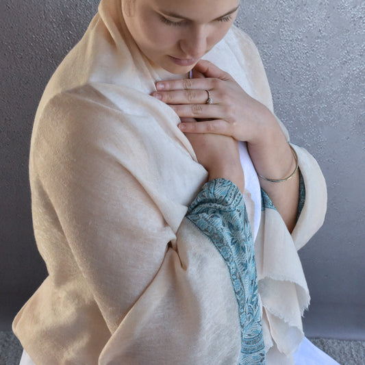 Woman wearing a beige shawl and white top with blue pattern, sitting against a gray wall.