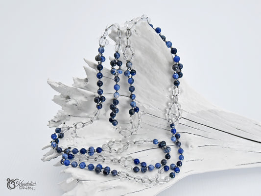 A sodalite and clear quartz Tantric necklace displayed on a white driftwood shell against a light gray background.