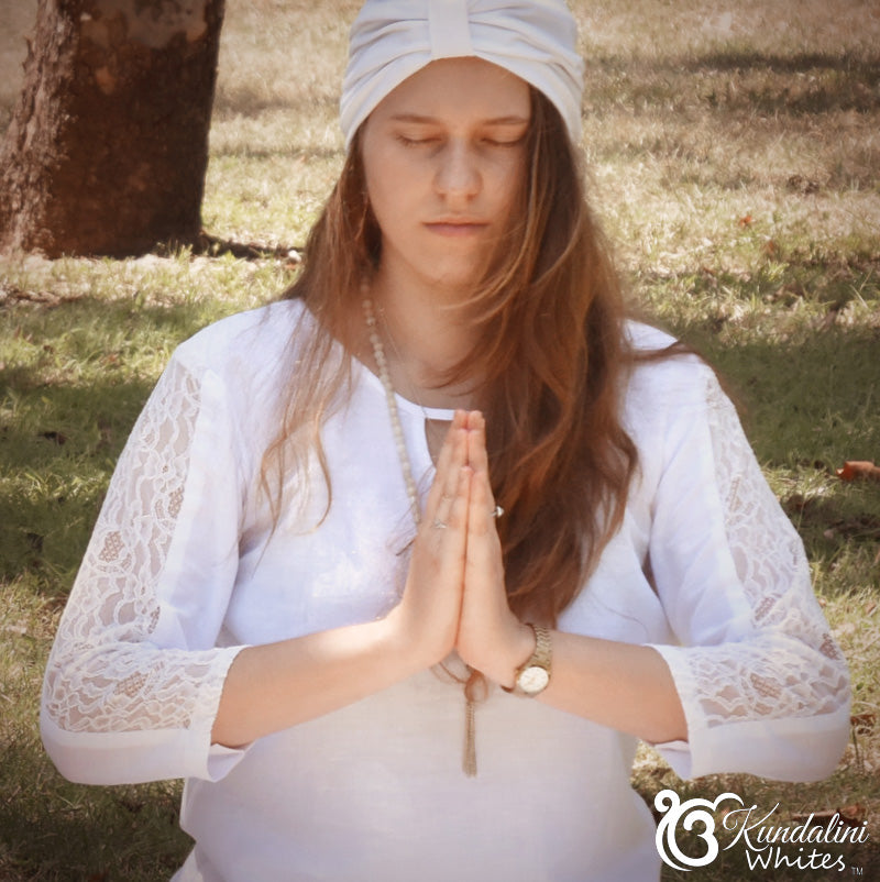 Woman in white traditional attire meditating outdoors with a tree and grass in the background.
