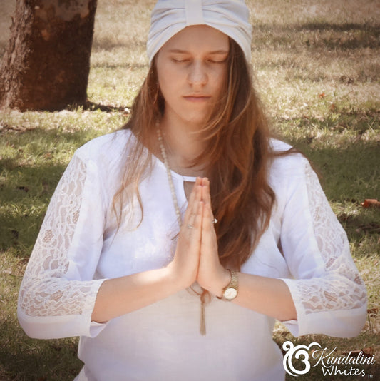 Woman in white traditional attire meditating outdoors with a tree and grass in the background.