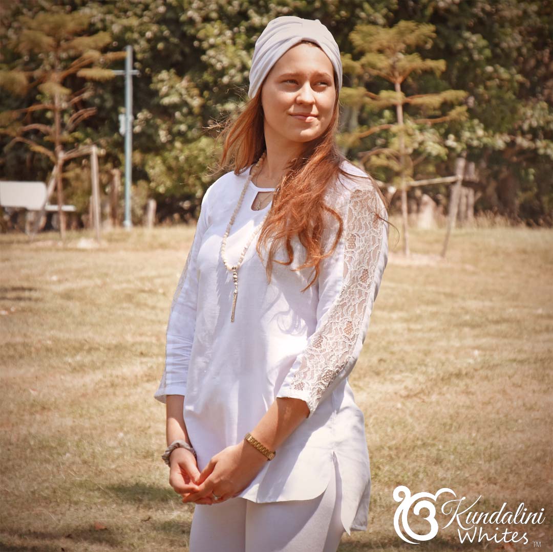 Woman in a white outfit with lace sleeves standing in a field, with 'Kundalini Whites' logo in the corner.