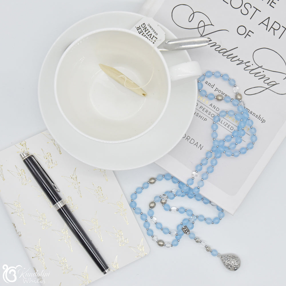 White cup with pen, light blue beaded necklace, and notebook on a white surface.