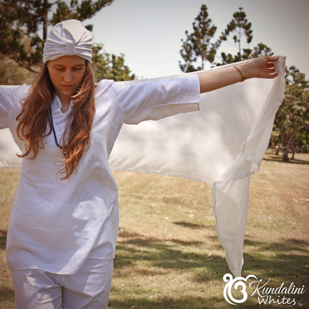 Person wearing a white outfit with a headscarf, standing outdoors with trees in the background.