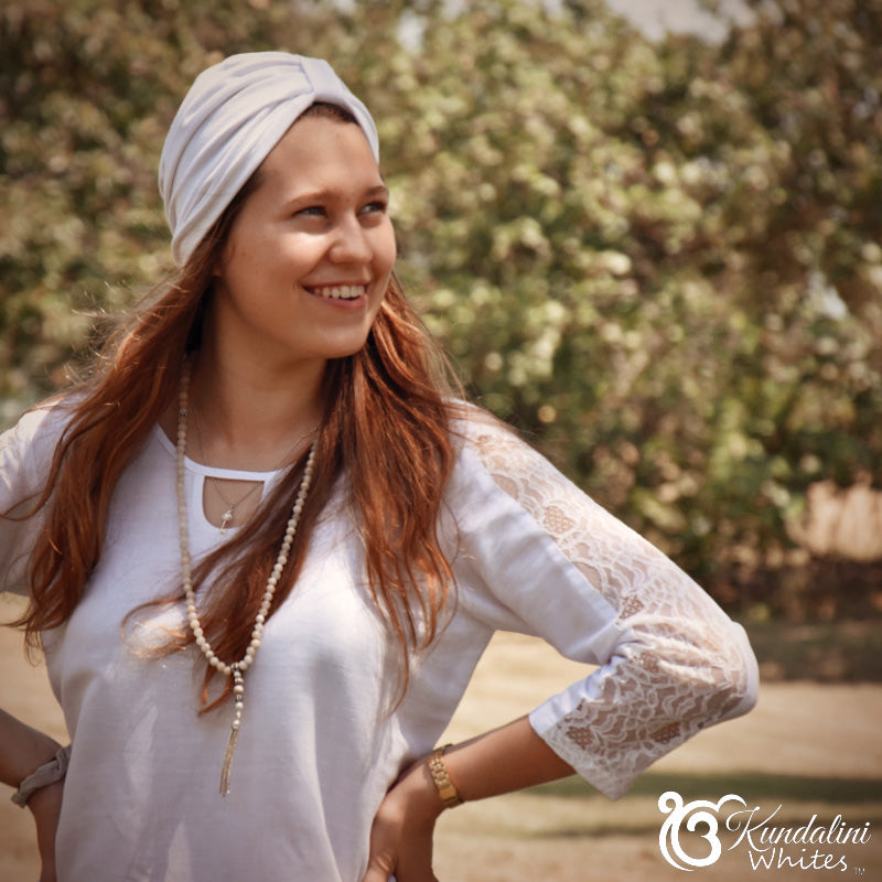Woman wearing a white top with lace sleeves and a headscarf, standing outdoors with greenery in the background.