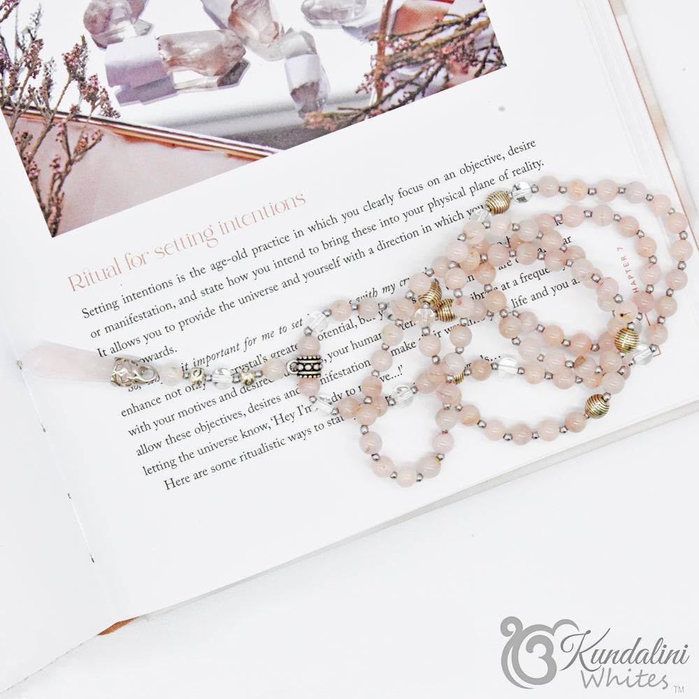Rose quartz mala necklace on an open book with 'Kundalini Whites' branding.