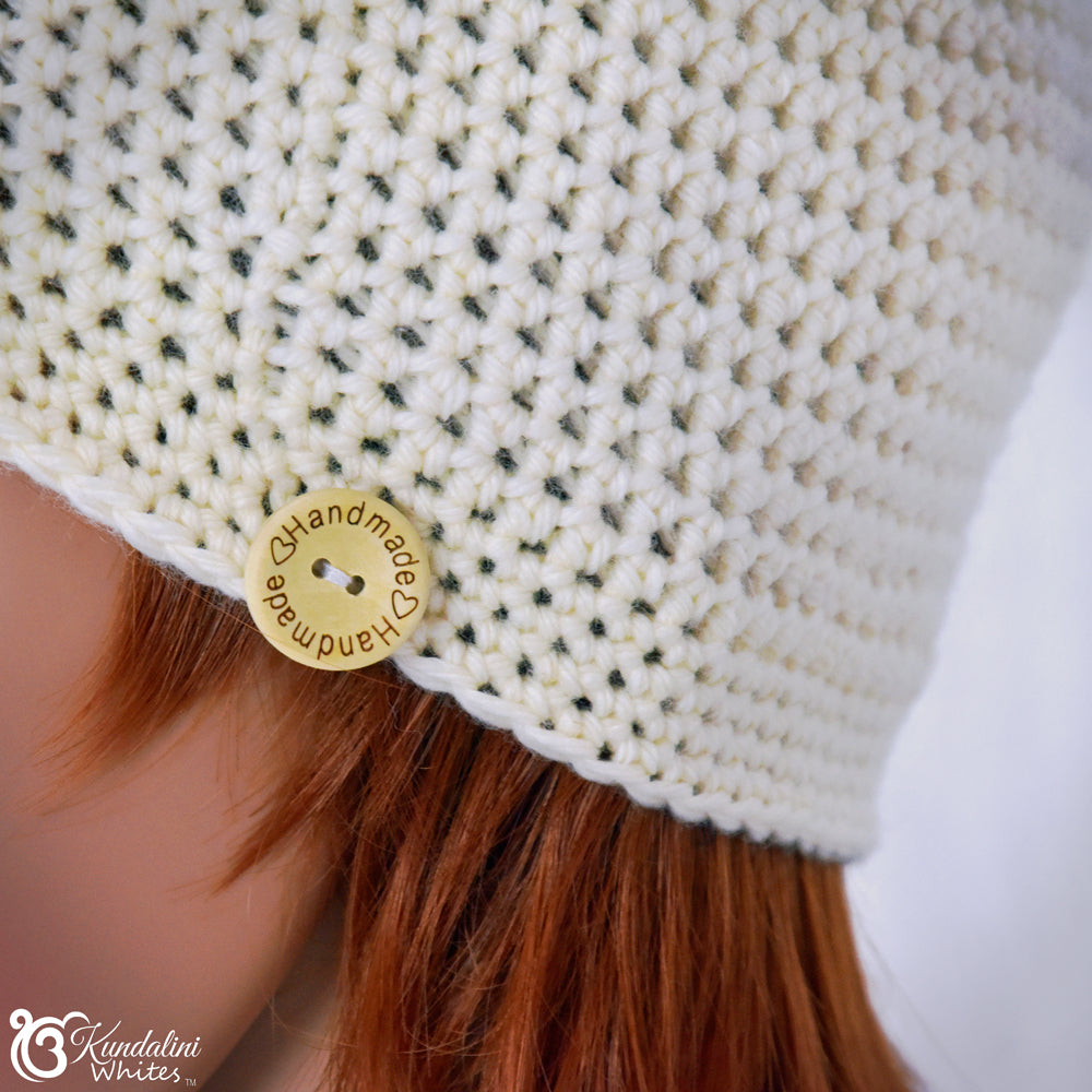 Close-up of a white knitted hat with a gold 'Handmade' button, worn by a person with red hair.