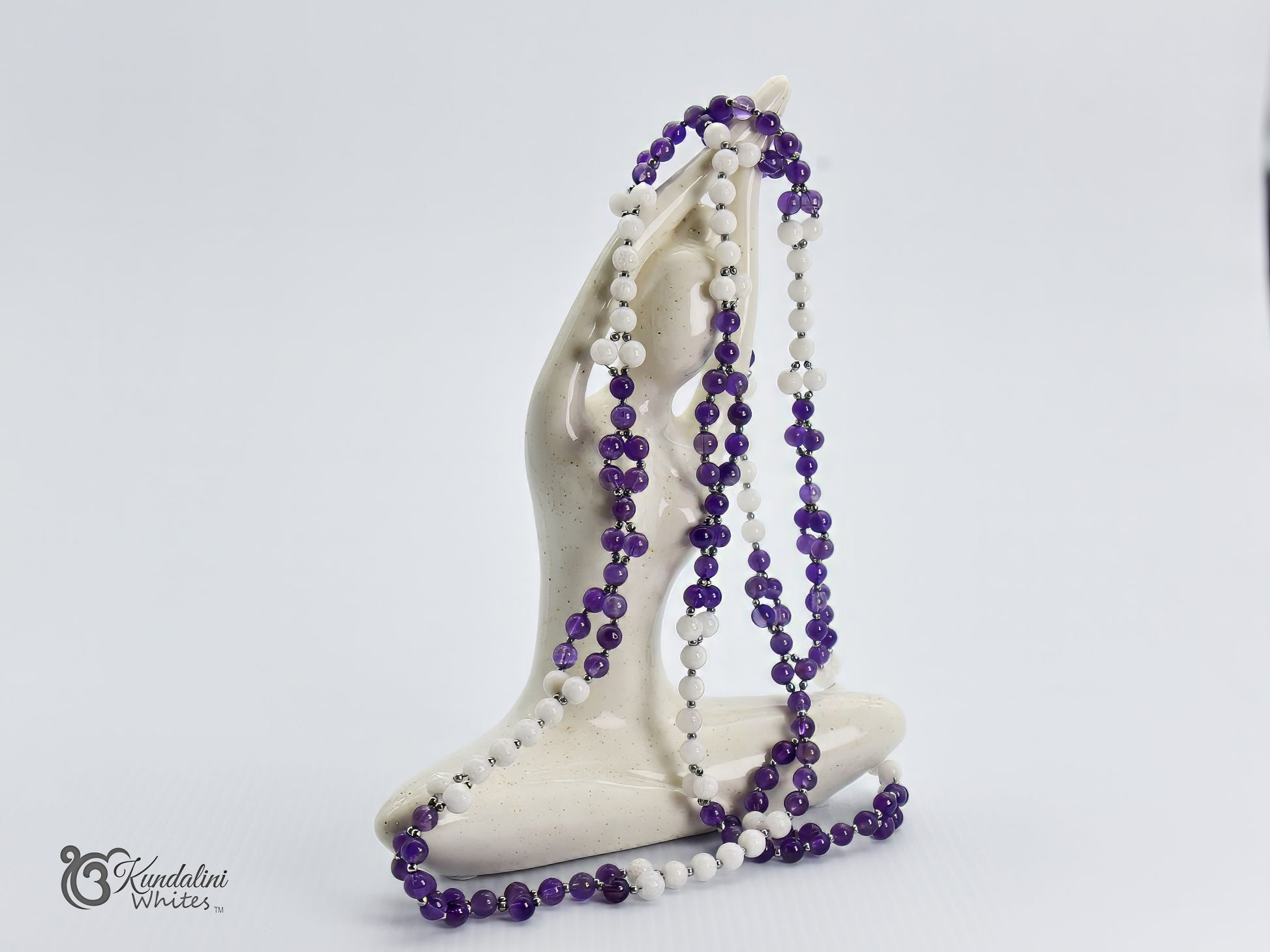 Purple and white beaded tantric necklace draped over a white sculpture on a light gray background.