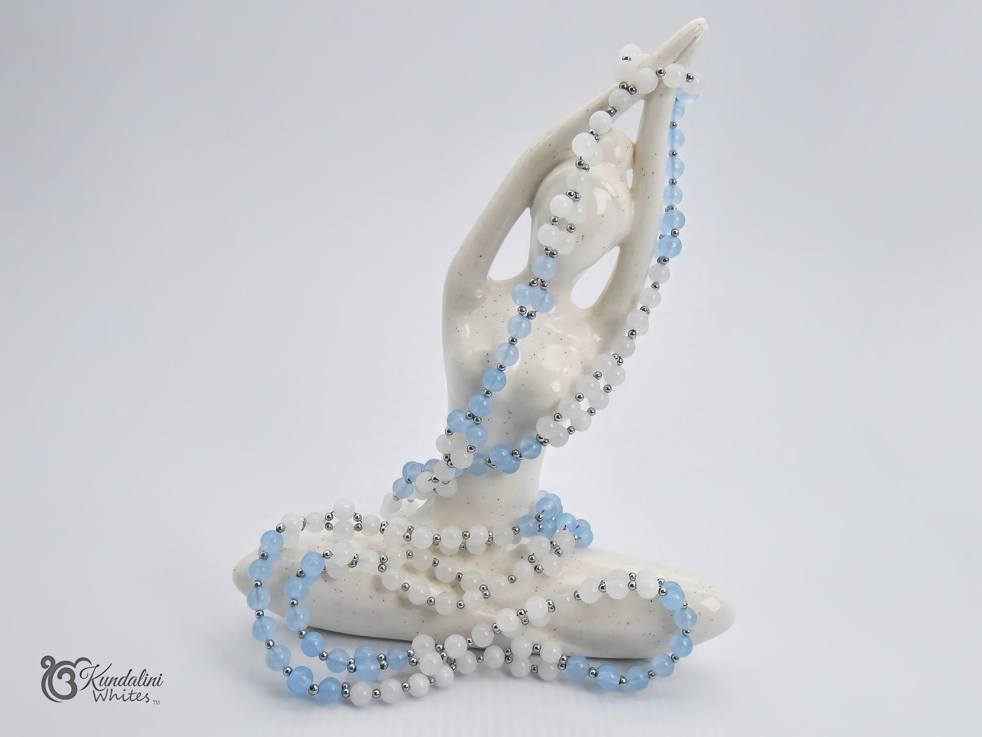 A white ceramic statue in a seated meditation pose, draped with an aquamarine and white jade Tantric Mala, on a light gray background.