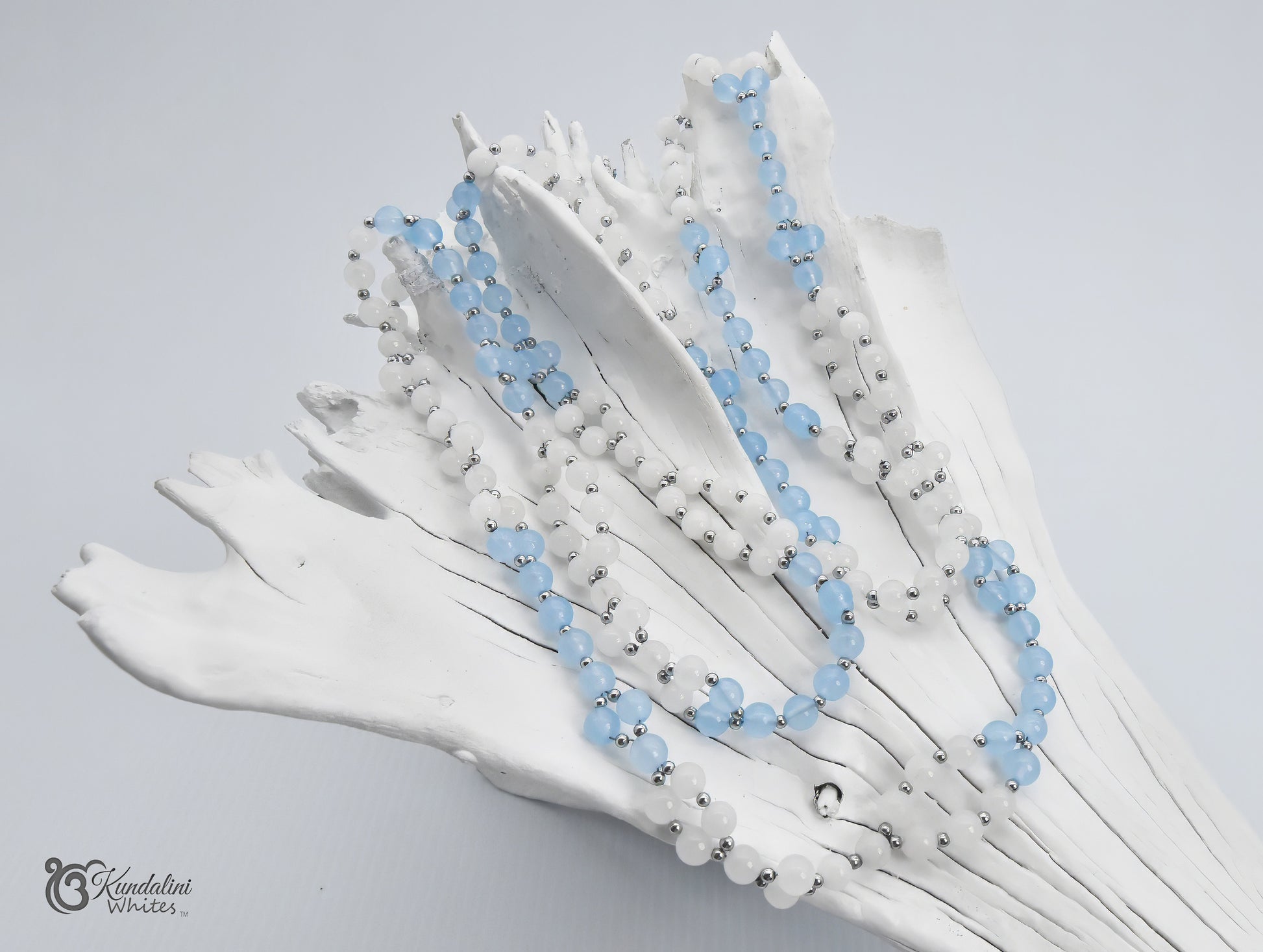 An aquamarine and white jade Tantric Mala draped over a white decorative branch or driftwood piece against a light gray background. 