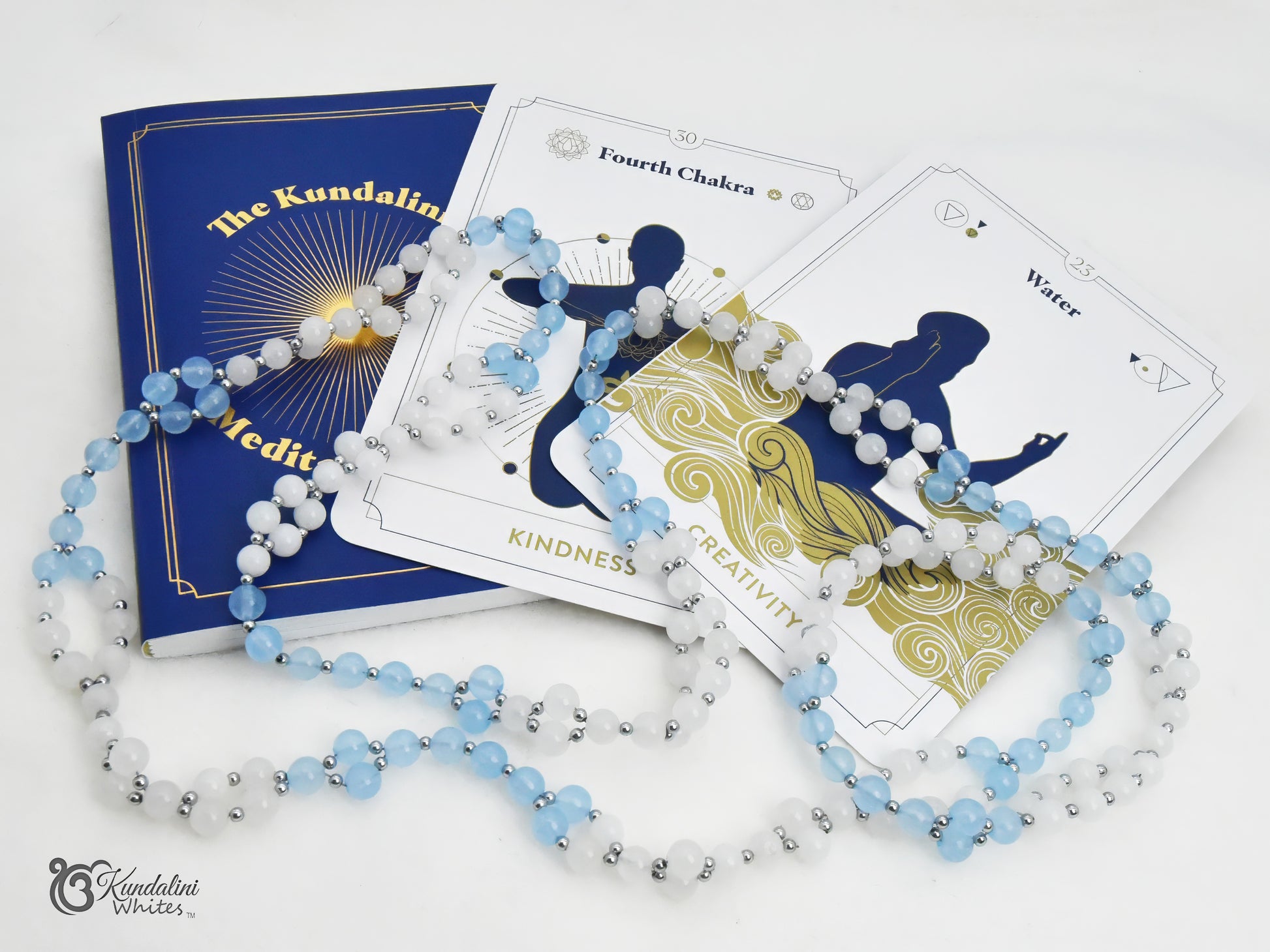 An aquamarine and white jade Tantric Mala arranged over a blue 'The Kundalini Meditation' book and a set of chakra cards on a white background.