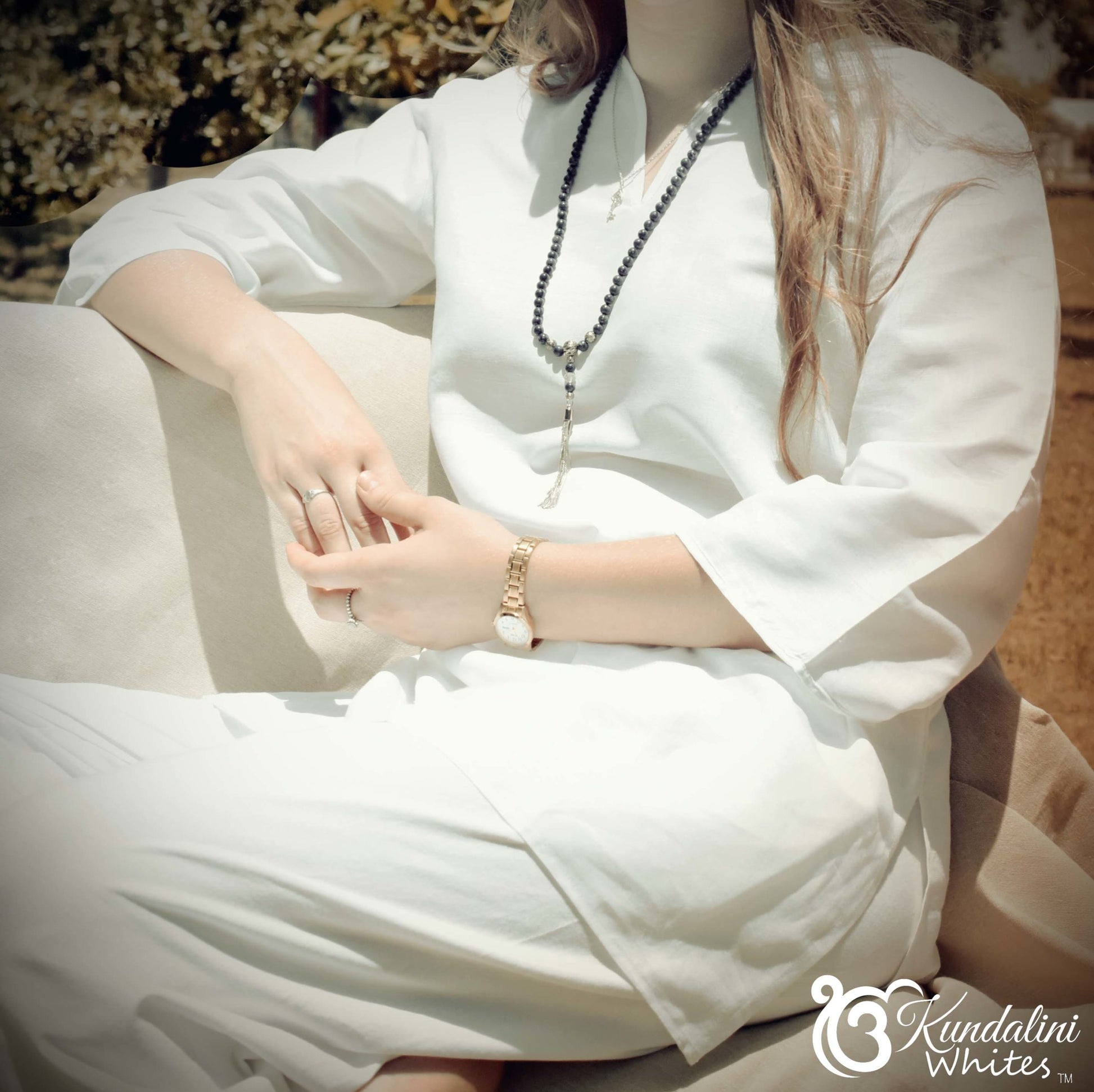 Person wearing a white dress with a necklace and bracelet, sitting on a stone surface.