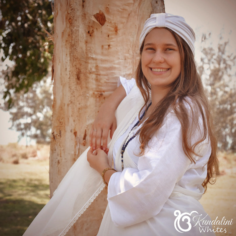 Woman in a white dress and headscarf standing next to a tree, with 'Kundalini Whites' logo in the corner.