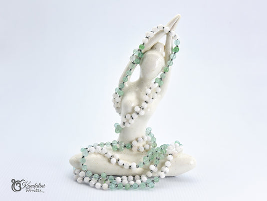 A white ceramic figurine of a seated woman with her hands raised, adorned with a fluorite and white agate Tantric Mala, set against a plain white background.
