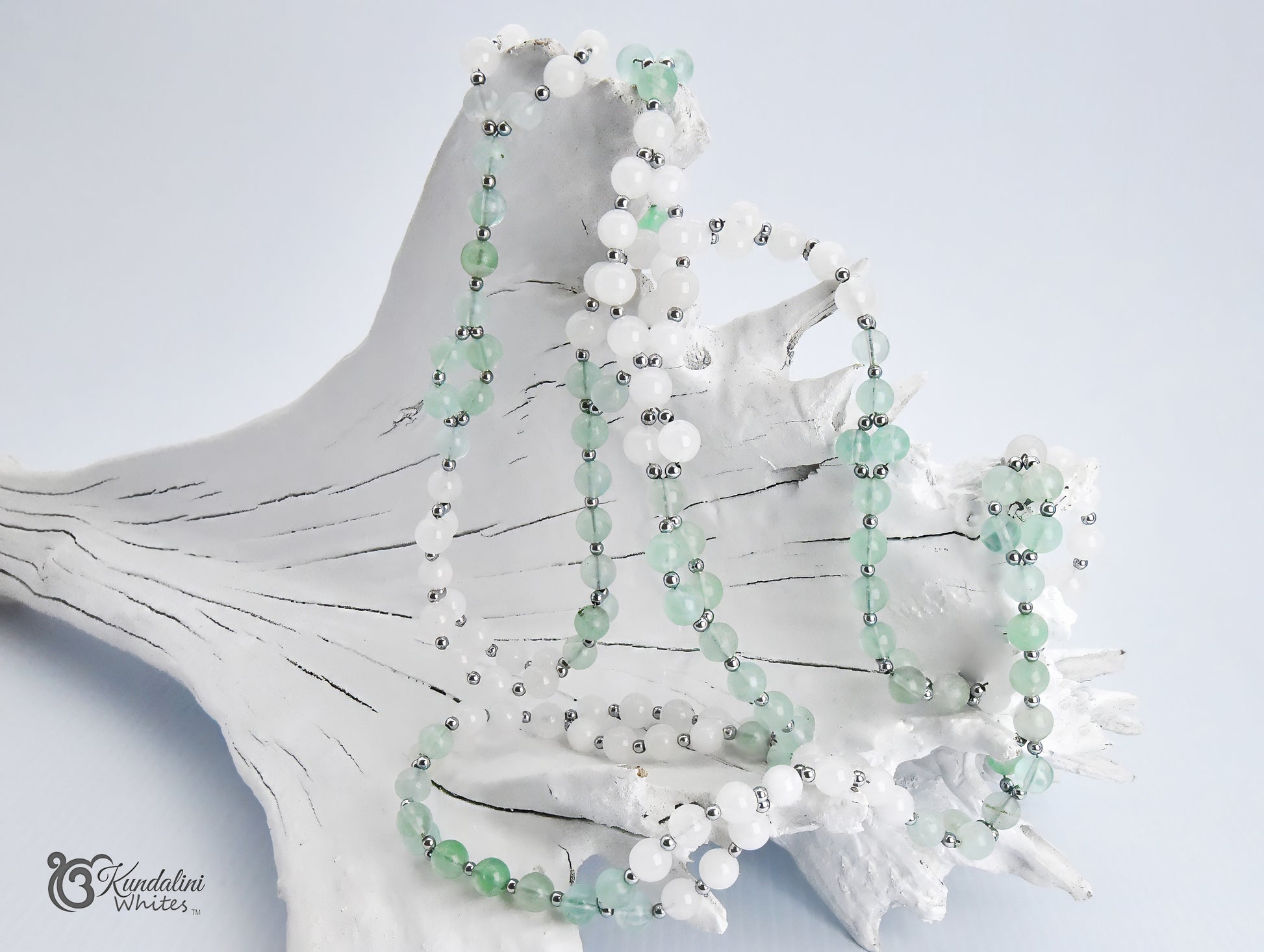 A fluorite and white jade Tantric Mala displayed on a white driftwood shell against a pale blue background. 