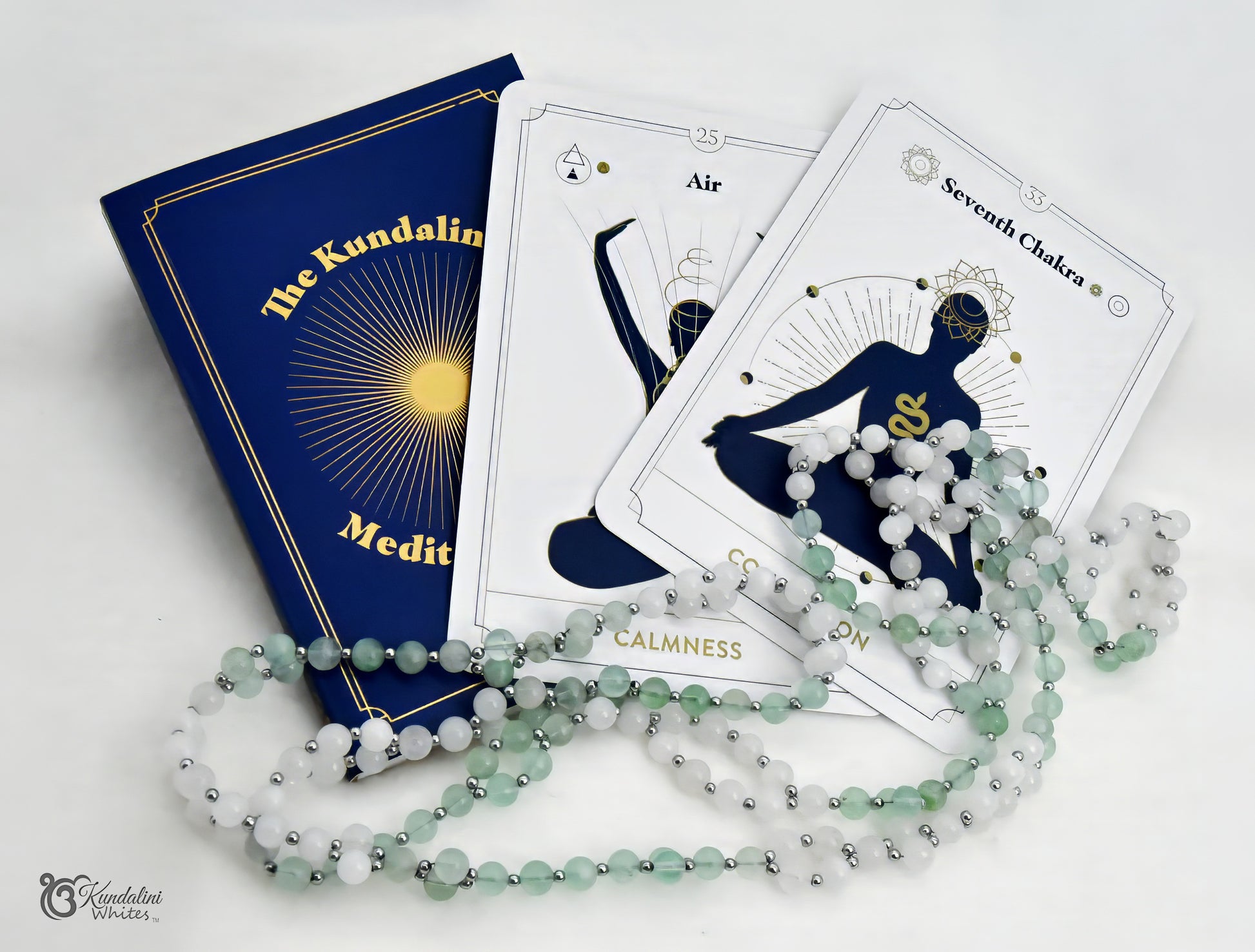 The Kundalini Meditation set featuring a deep blue book with gold lettering, white tarot-style cards with gold illustrations, and a fluorite and white jade Tantric Mala, all arranged on a white background.