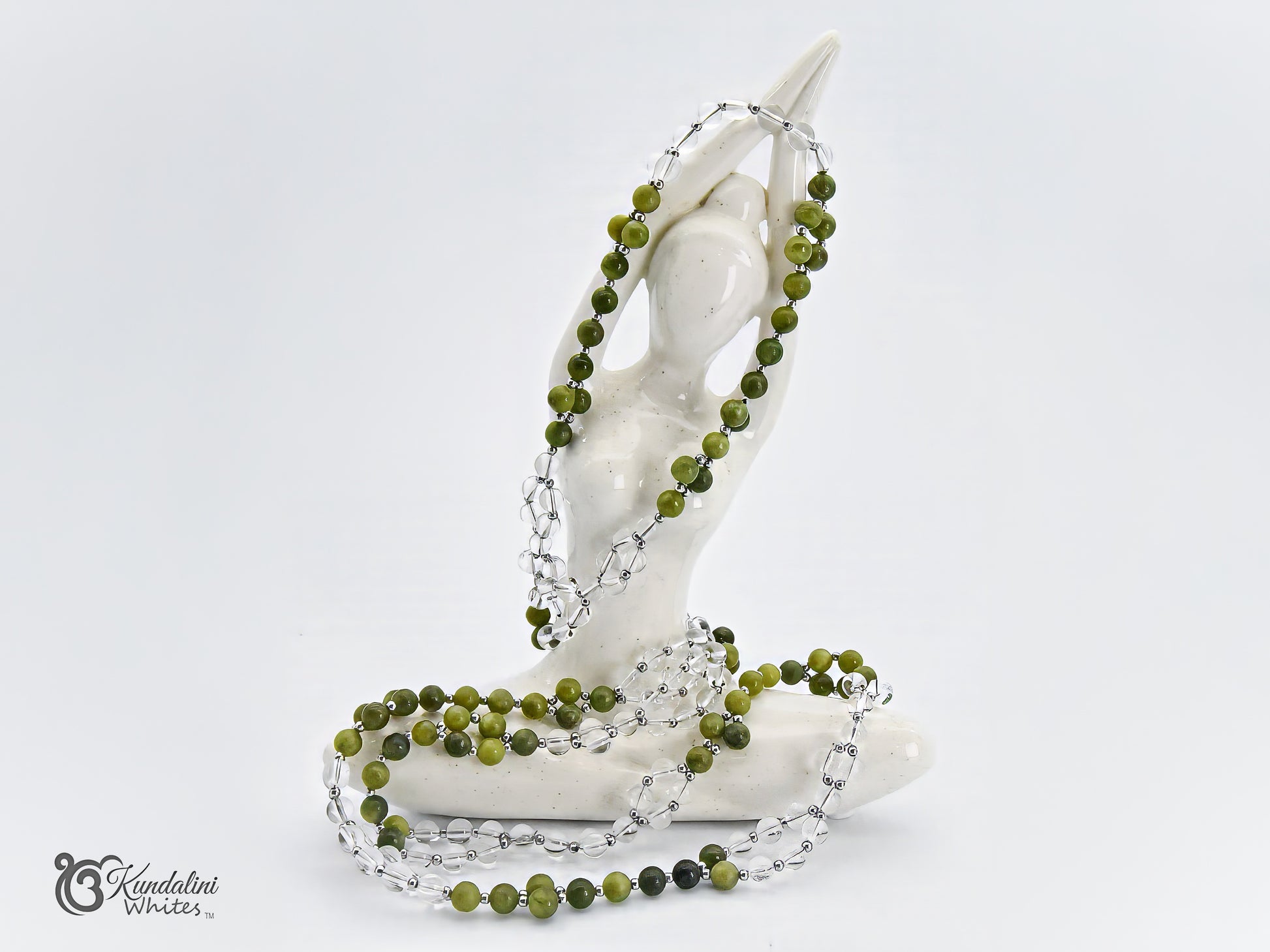 A Tantric mala with green jade and clear quartz beads draped over a white ceramic figurine, photographed against a seamless white background.