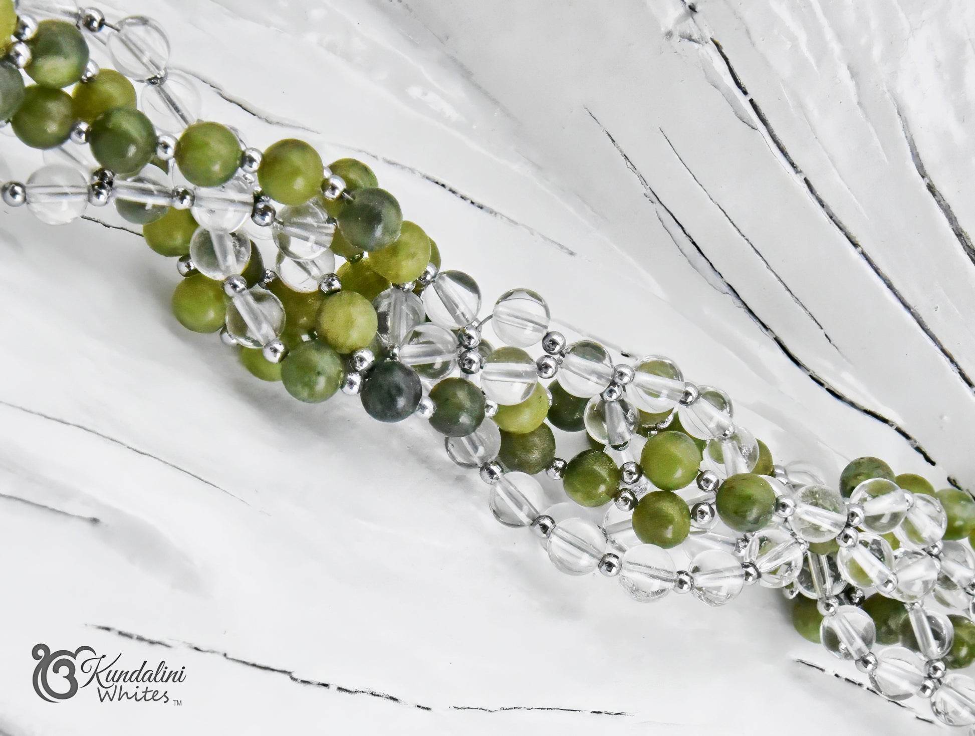 A close-up of a green jade and clear quartz Tantric Mala with beads separated by small silver spacer beads, laid diagonally on a white textured background. The brand name 'Kundalini Whites' is visible in the bottom-left corner.