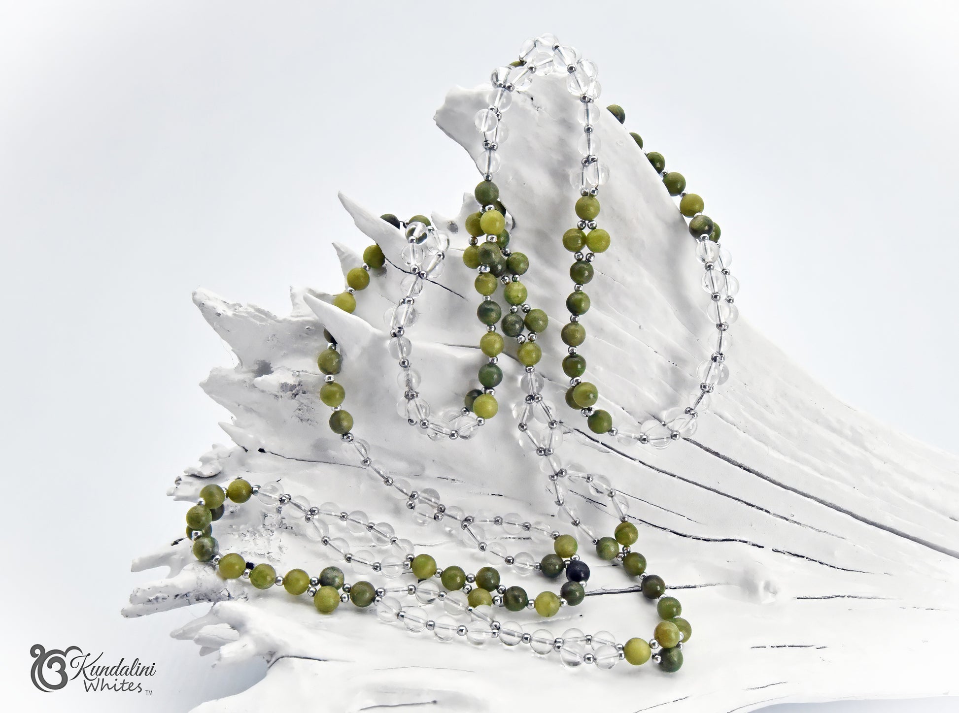 A Tantric Mala with green jade and clear quartz beads is draped over a white decorative driftwood piece on a clean white background. A watermark reading 'Kundalini Whites' is visible in the lower-left corner.