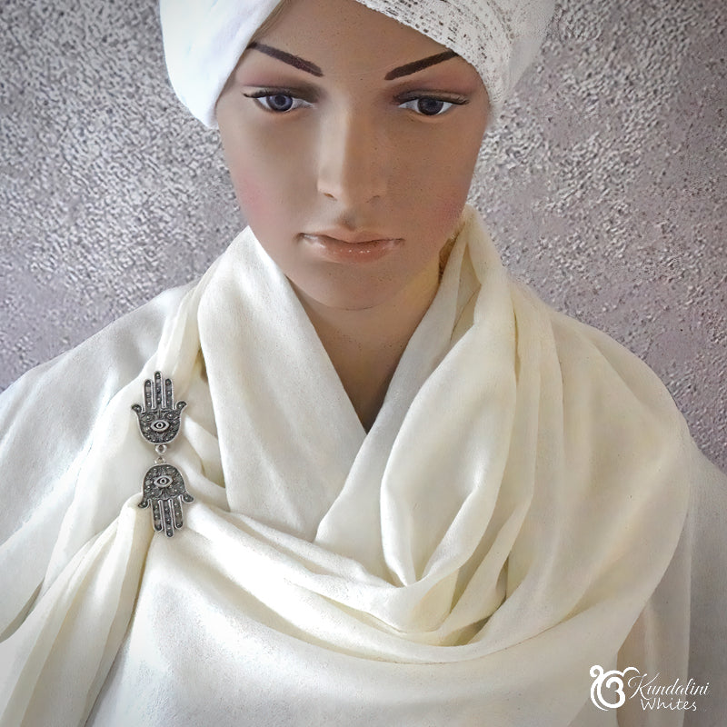 Mannequin wearing a white shawl with a Hamsa hand pendant against a textured gray wall.