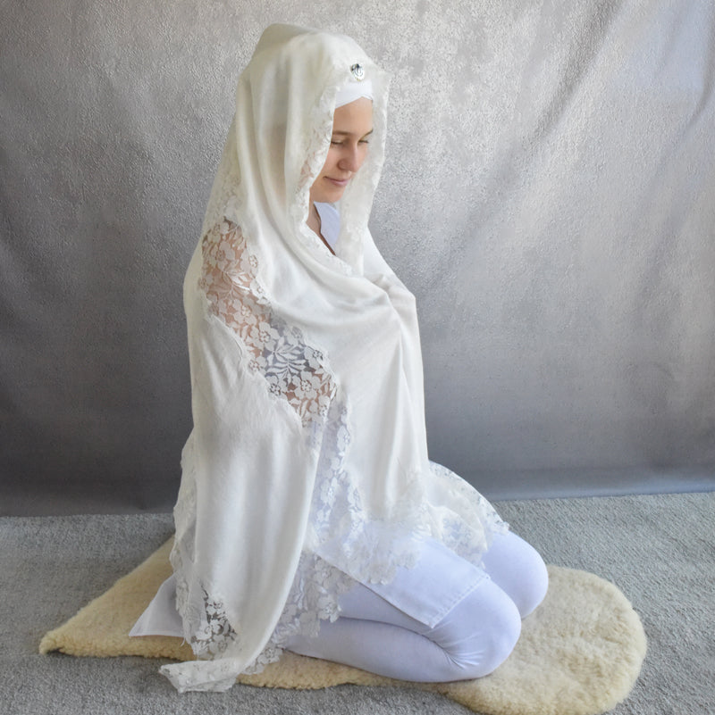 Woman wearing a white shawl with lace details, sitting on a rug against a plain background