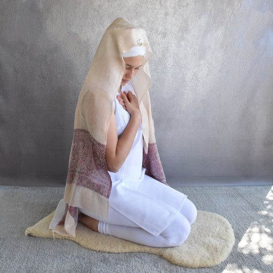 Woman in white outfit with beige shawl kneeling on a mat against a plain background