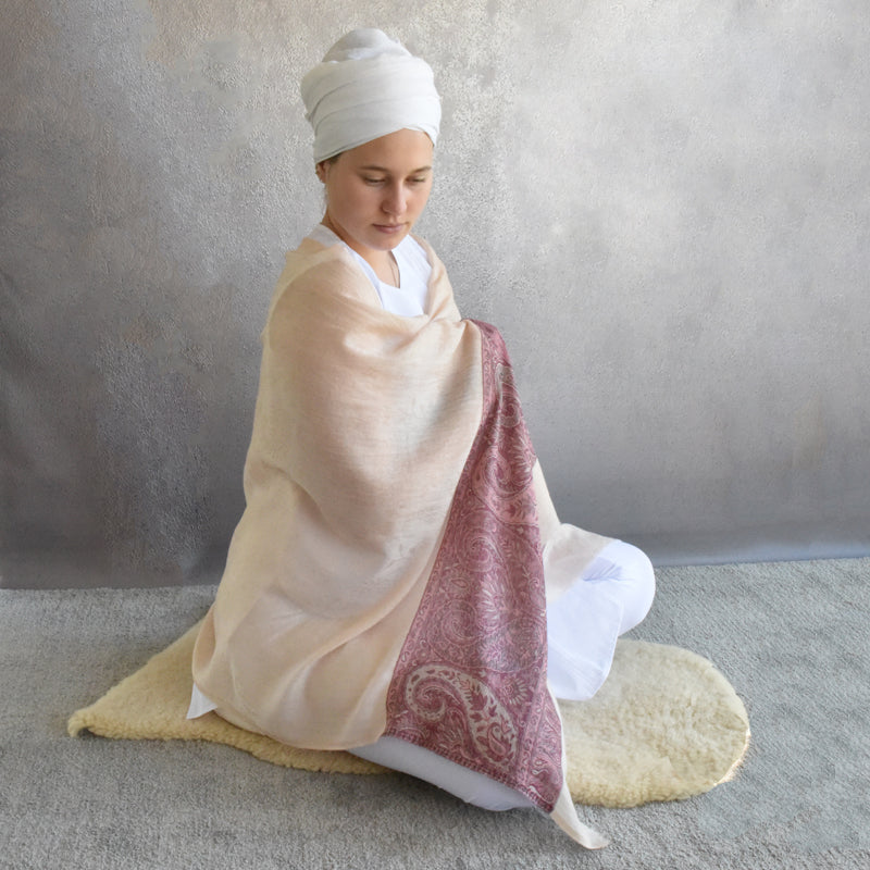 Person wrapped in a beige shawl with a red pattern, sitting on a rug against a plain wall.