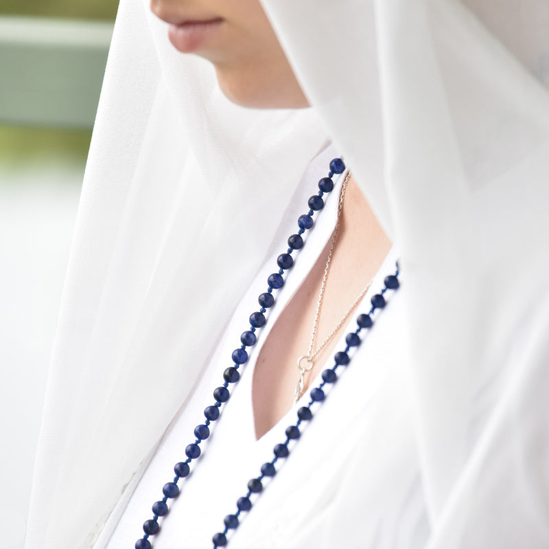 Person wearing a white garment with a blue beaded necklace and gold chain.