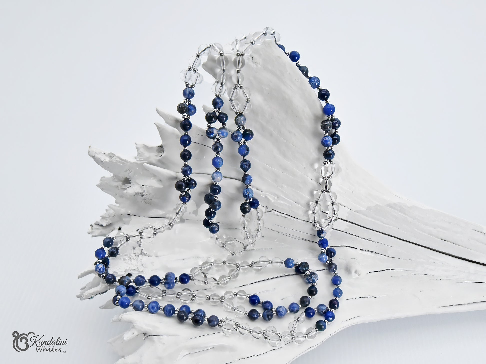 A sodalite and clear quartz Tantric necklace displayed on a white driftwood shell against a light gray background. 