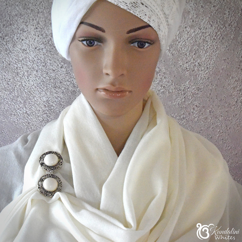 Mannequin head wearing a white scarf with decorative brooches against a textured gray wall.